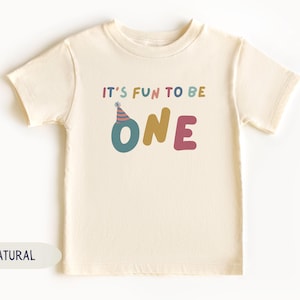 May include: A cream-coloured toddler t-shirt with the text "IT'S FUN TO BE ONE" in colourful letters. The word "ONE" is in teal, gold, and pink. A party hat graphic sits above the letter "O".