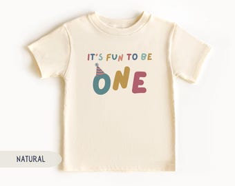 May include: A cream-colored toddler t-shirt with the text "IT'S FUN TO BE ONE" in colorful letters. The word "ONE" is in teal, gold, and pink. A party hat graphic sits above the letter "O".