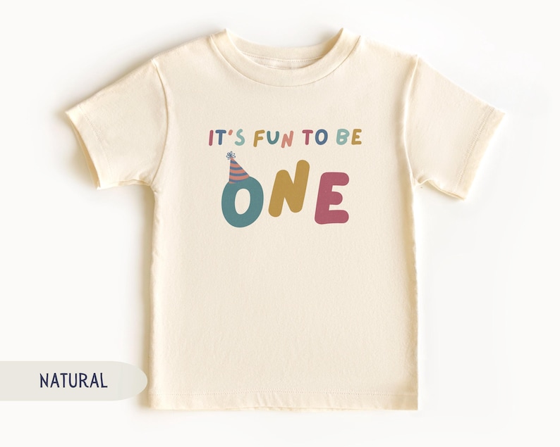 May include: A cream-coloured toddler t-shirt with the text "IT'S FUN TO BE ONE" in colourful letters. The word "ONE" is in teal, gold, and pink. A party hat graphic sits above the letter "O".