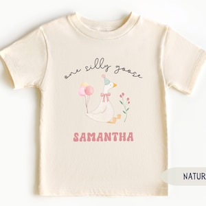 Pu&ograve; includere: Una maglietta bianca con una simpatica illustrazione di un'oca che indossa un cappello da festa e tiene dei palloncini. Il testo "one silly goose" &egrave; scritto sopra l'oca e il nome "SAMANTHA" &egrave; scritto sotto l'oca. La maglietta &egrave; realizzata in tessuto naturale.