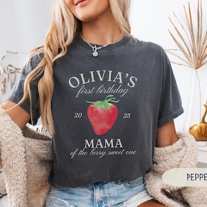 May include: Dark gray t-shirt with the text "OLIVIA'S first birthday" above a red strawberry graphic. Below the strawberry, the text reads "MAMA of the berry sweet one". The numbers "20" and "25" are on either side of the strawberry.