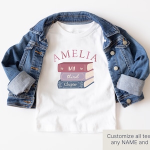 May include: A white t-shirt featuring the name "AMELIA" in pink script above a stack of three books. The books are pink, purple, and blue, with the words "MY", "third", and "Chapter". A denim jacket is draped over the shirt.