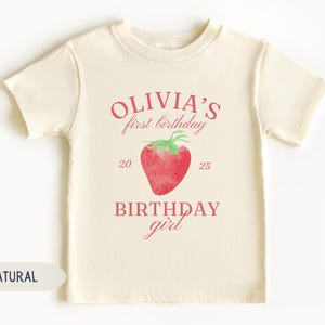 May include: A cream-colored t-shirt with the text "Olivia's first birthday" in pink, along with a watercolor strawberry illustration. The text "BIRTHDAY girl" is also printed on the shirt.