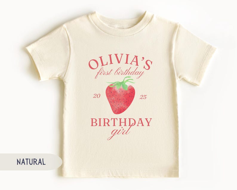 May include: A cream-colored t-shirt with the text "Olivia's first birthday" in pink, along with a watercolor strawberry illustration. The text "BIRTHDAY girl" is also printed on the shirt.
