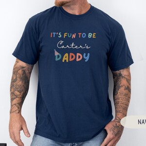 May include: A navy blue t-shirt featuring the phrase "IT'S FUN TO BE Carter's DADDY" in a colourful, playful font. The design includes a party hat graphic. The t-shirt has a crew neck and short sleeves, suitable for casual wear.
