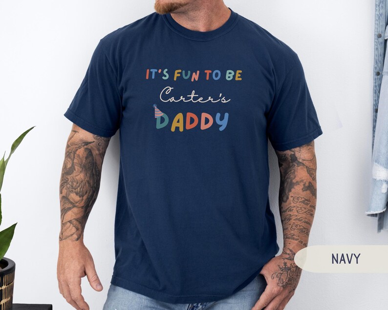 May include: A navy blue t-shirt featuring the phrase "IT'S FUN TO BE Carter's DADDY" in a colourful, playful font. The design includes a party hat graphic. The t-shirt has a crew neck and short sleeves, suitable for casual wear.