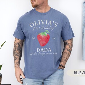 May include: Blue jean-colored t-shirt with the text "OLIVIA'S first birthday" above a red strawberry graphic. Below the strawberry, the text reads "DADA of the berry sweet one". The numbers "20" and "25" are on either side of the strawberry.