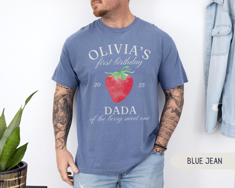 May include: Blue jean-colored t-shirt with the text "OLIVIA'S first birthday" above a red strawberry graphic. Below the strawberry, the text reads "DADA of the berry sweet one". The numbers "20" and "25" are on either side of the strawberry.