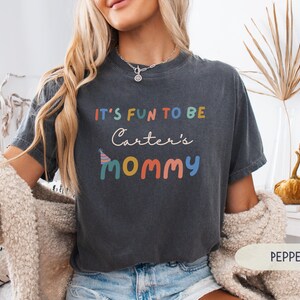 May include: A dark grey t-shirt with the text "IT'S FUN TO BE Carter's MOMMY" in colourful letters. The shirt has a relaxed fit and is made of soft material. The shirt is a casual wear item.