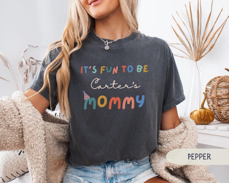 May include: A dark grey t-shirt with the text "IT'S FUN TO BE Carter's MOMMY" in colourful letters. The shirt has a relaxed fit and is made of soft material. The shirt is a casual wear item.