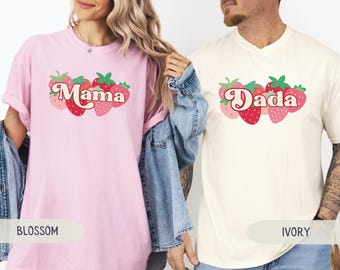 May include: Two t-shirts, one blossom pink and one ivory, each with a strawberry design. The pink shirt says "Mama" and the ivory shirt says "Dada". The strawberries are red and pink with green leaves.