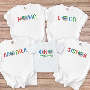 May include: White t-shirts with colorful text and book graphics. The shirts say "MAMA", "DADA", "BROTHER", "SISTER", and "ONE for the books". The text and book graphics are in various colors.