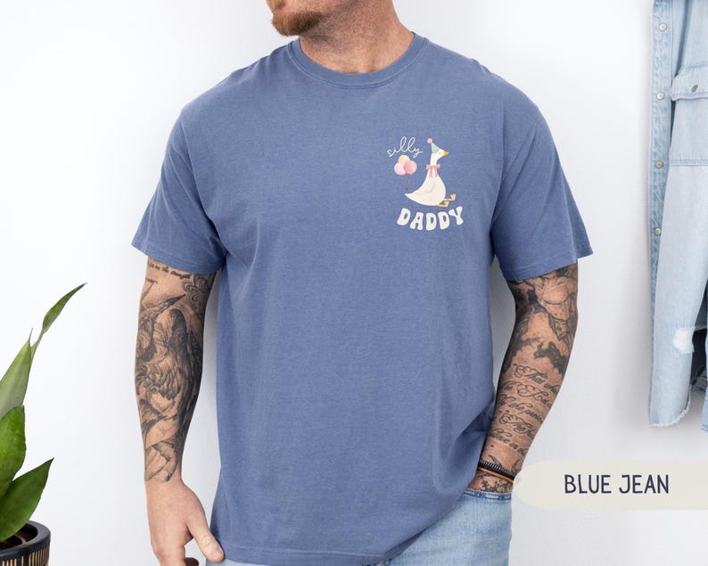 Pu&ograve; includere: T-shirt in jeans blu con un'oca di cartone animato che indossa un cappello da festa e tiene dei palloncini. Il testo "Silly Daddy" &egrave; stampato sulla maglietta.