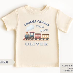 May include: A cream-colored toddler t-shirt with a train graphic. The design includes the text "CHUGGA CHUGGA TWO TWO" above a train illustration and the name "OLIVER" below. The shirt is made of soft material.