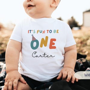 May include: White t-shirt with the text "IT'S FUN TO BE ONE" in colourful letters. The word "ONE" is in large, bold letters. Below the text is the name "Carter" in a cursive font. The shirt has a birthday party theme.