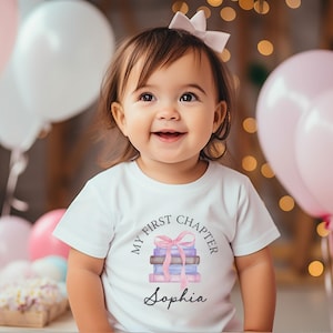 Może przedstawiać: Biała koszulka z napisem "MY FIRST CHAPTER" i imieniem "Sophia". Koszulka przedstawia akwarelową ilustrację stosu książek związanego różową kokardą. Obraz zawiera różowe i białe balony.