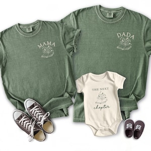 Könnte beinhalten: Olivgrüne T-Shirts mit den Aufdrucken "MAMA" und "DADA", sowie ein Baby-Body mit "THE NEXT CHAPTER". Jedes Kleidungsstück hat ein Buch- und Blumendesign. Das Bild enthält auch ein Paar Turnschuhe und Babyschuhe.