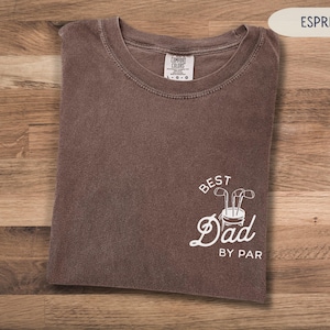 May include: A brown t-shirt with the text "Best Dad By Par" in white. The text is accompanied by a graphic of a golf bag with golf clubs.