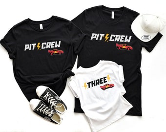 Könnte beinhalten: Drei schwarze T-Shirts mit einem Rennauto-Motiv. Die Erwachsenen-Shirts sagen "Pit Crew" mit einem Blitz und einem roten Rennwagen. Das Kinder-Shirt sagt "Three" mit einem Blitz und einem roten Rennwagen.
