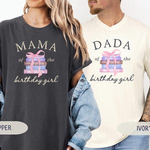 May include: Two t-shirts, one pepper-colored and one ivory-colored, each with a graphic. The pepper shirt says "MAMA of the birthday girl" and the ivory shirt says "DADA of the birthday girl". Both shirts feature a stack of books tied with a pink bow.