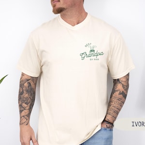 May include: A cream colored t-shirt with a green and white golf-themed graphic that says "Best Grandpa by Par".