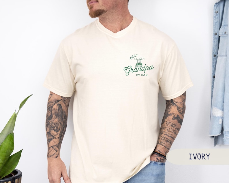 May include: A cream colored t-shirt with a green and white golf-themed graphic that says "Best Grandpa by Par".