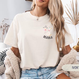 Pu&ograve; includere: Una t-shirt bianca con una grafica di un'oca con il testo "silly mama". La modella indossa la t-shirt con jeans blu e un cardigan color crema. Lo sfondo &egrave; una superficie di legno chiaro.