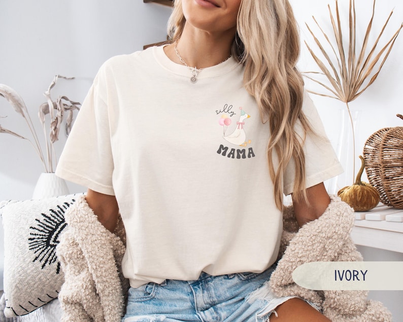 Pu&ograve; includere: Una t-shirt bianca con una grafica di un'oca con il testo "silly mama". La modella indossa la t-shirt con jeans blu e un cardigan color crema. Lo sfondo &egrave; una superficie di legno chiaro.