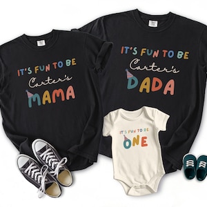 May include: Black t-shirts and a white babygrow with the text "IT'S FUN TO BE" and "Carter's" followed by "MAMA", "DADA", and "ONE" in colourful letters. A pair of black trainers and blue baby shoes are also in the image.