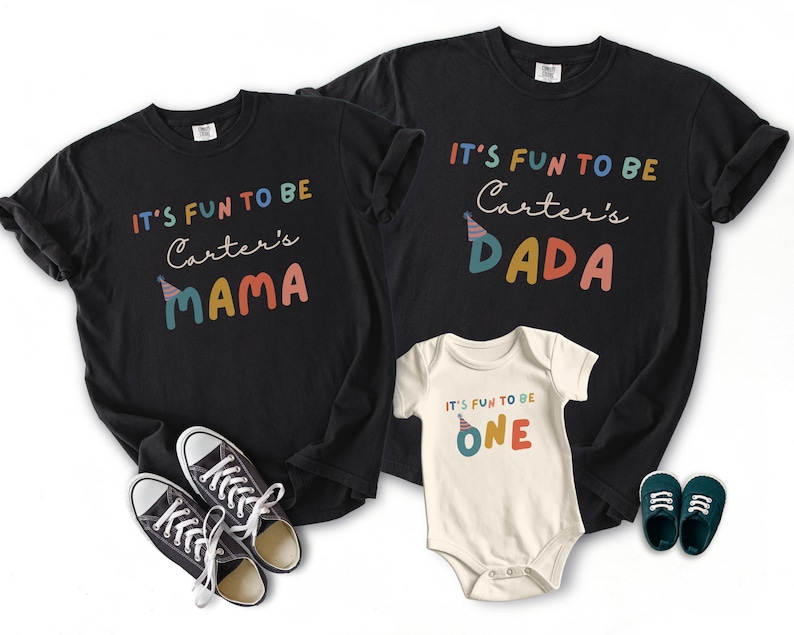 May include: Black t-shirts and a white babygrow with the text "IT'S FUN TO BE" and "Carter's" followed by "MAMA", "DADA", and "ONE" in colourful letters. A pair of black trainers and blue baby shoes are also in the image.
