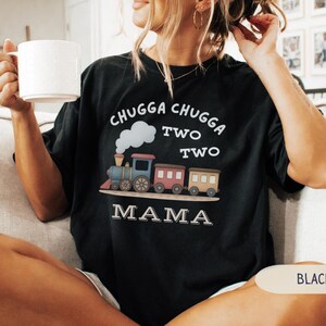 May include: Black t-shirt with a colorful train graphic. The design includes the words "CHUGGA CHUGGA TWO TWO TWO MAMA" in white. The train features blue, pink, and brown cars with a white cloud and smoke detail.
