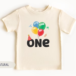 K&ouml;nnte beinhalten: Cremefarbenes Kleinkind-T-Shirt mit einer bunten Ballon-Grafik. Die Ballons sind blau, gr&uuml;n, gelb und rot, mit den Worten "IT'S FUN TO BE" darauf. Unter den Ballons steht das Wort "ONE" in gro&szlig;en schwarzen Buchstaben, mit einer Partyh&uuml;tchen &uuml;ber dem "O".
