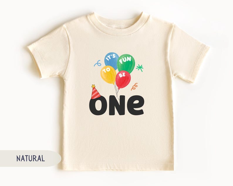 K&ouml;nnte beinhalten: Cremefarbenes Kleinkind-T-Shirt mit einer bunten Ballon-Grafik. Die Ballons sind blau, gr&uuml;n, gelb und rot, mit den Worten "IT'S FUN TO BE" darauf. Unter den Ballons steht das Wort "ONE" in gro&szlig;en schwarzen Buchstaben, mit einer Partyh&uuml;tchen &uuml;ber dem "O".