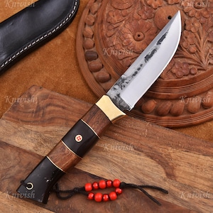 Può includere: Un coltello artigianale con una lama in acciaio lucido e un manico in legno. Il manico presenta dettagli in ottone e una sezione nera con un emblema rosso. Un fodero in pelle è visibile sullo sfondo. Il coltello è lungo circa 23 cm.