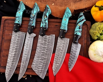 May include: A collection of six kitchen knives with patterned blades and turquoise and black handles. The blades have a distinctive design, and the handles feature gold-coloured rivets. The knives are displayed on a wooden chopping board.