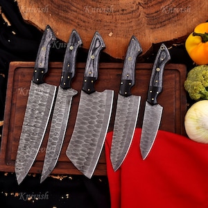 May include: A set of six Damascus steel kitchen knives with wooden handles. The blades have a unique pattern, and the handles are dark gray with brass accents. The knives are arranged on a wooden cutting board, with vegetables in the background.