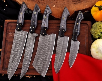 May include: A set of six Damascus steel kitchen knives with wooden handles. The blades have a unique pattern, and the handles are dark gray with brass accents. The knives are arranged on a wooden cutting board, with vegetables in the background.