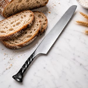 May include: A serrated bread knife with a long, silver-colored metal blade and a dark, twisted metal handle. The knife is positioned diagonally next to several slices of crusty, light brown bread on a white surface with scattered flour.