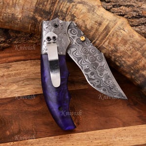 May include: A folding knife with a patterned steel blade and a purple handle. The blade has a unique swirling pattern. The handle is a deep purple color. The knife has a silver pocket clip and a gold-colored rivet.