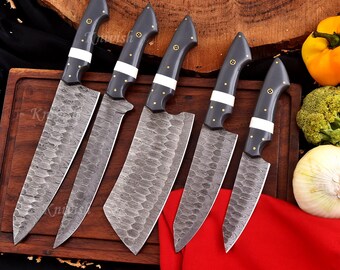 May include: A collection of five kitchen knives featuring dark grey handles, white detailing, and patterned blades. The handles are embellished with gold-toned rivets. The knives are displayed on a wooden chopping board, with vegetables and a red cloth in the background.