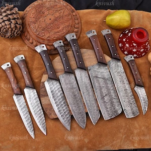 May include: A collection of eight handcrafted knives with dark wooden handles and patterned steel blades. The blades vary in shape and size, with some featuring a textured surface. The knives are arranged on a brown surface.