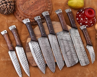 May include: A collection of eight handcrafted knives with dark wooden handles and patterned steel blades. The blades vary in shape and size, with some featuring a textured surface. The knives are arranged on a brown surface.