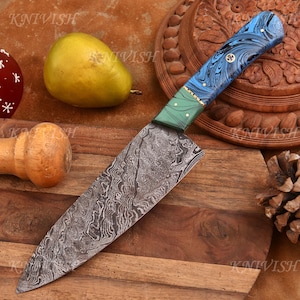 May include: A Damascus steel chef's knife with a patterned blade and a handle featuring blue, green, and gold accents. The knife rests on a wooden cutting board, with a pear and other decorative items in the background.