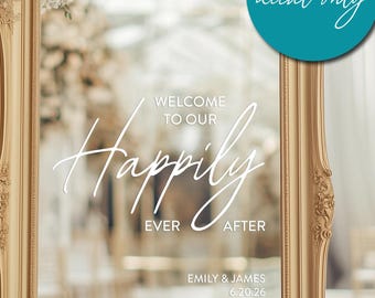 May include: A gold-framed mirror with a white vinyl decal that reads "Welcome to our Happily Ever After" with the names Emily & James and the date 6.20.26. The decal is for a wedding reception.