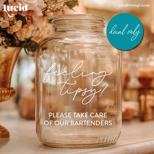 May include: A clear glass jar with a wide mouth, featuring the text "feeling tipsy?" and "PLEASE TAKE CARE OF OUR BARTENDERS" in white script. A teal circle with the words "decal only" is visible. The jar is likely for a bar or event.