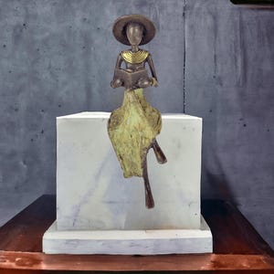 Könnte beinhalten: Eine Bronzeskulptur einer Person, die einen Hut und einen langen Rock trägt, auf einem weißen Sockel sitzt und ein Buch liest. Die Skulptur hat eine goldene Halskette und ist in Grün- und Brauntönen bemalt.