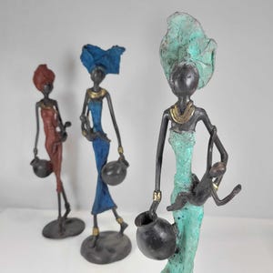 Puede incluir: Tres figuritas decorativas de mujeres con vestimenta tradicional africana. Las mujeres sostienen ollas y una de ellas lleva un niño. Las figuritas están hechas de metal y pintadas en tonos azules, verdes y rojos.