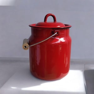 Puede incluir: Un cántaro de leche de esmalte rojo brillante con una tapa a juego que presenta un asa central. El recipiente incluye un asa metálica abatible con un agarre cilíndrico de madera en un lado. La superficie es lisa y brillante con un borde negro.