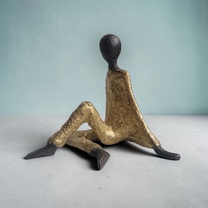 Könnte beinhalten: Eine Skulptur einer sitzenden Figur mit einem glatten, dunkel anthrazitfarbenen Kopf und Gliedmaßen, kontrastiert durch einen strukturierten, metallisch goldfarbenen Torso und Beine. Die Figur ist in einer entspannten, liegenden Pose dargestellt.