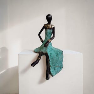 Könnte beinhalten: Bronzefarbene Skulptur einer sitzenden Figur mit türkisfarbenem Kleid und goldenen Akzenten. Die Figur posiert mit einem ausgestreckten Bein und Armen auf dem Schoß. Die Skulptur steht auf einem weißen quadratischen Sockel.